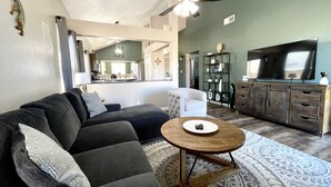 A living room with a dark-colored sofa, a round wooden coffee table, and a patterned rug. There is a television on a wooden cabinet and a ceiling fan above. - ABQ Home Overlooking Mountain- Near Sandia Tramway + Balloon Fiesta  (Albuquerque)