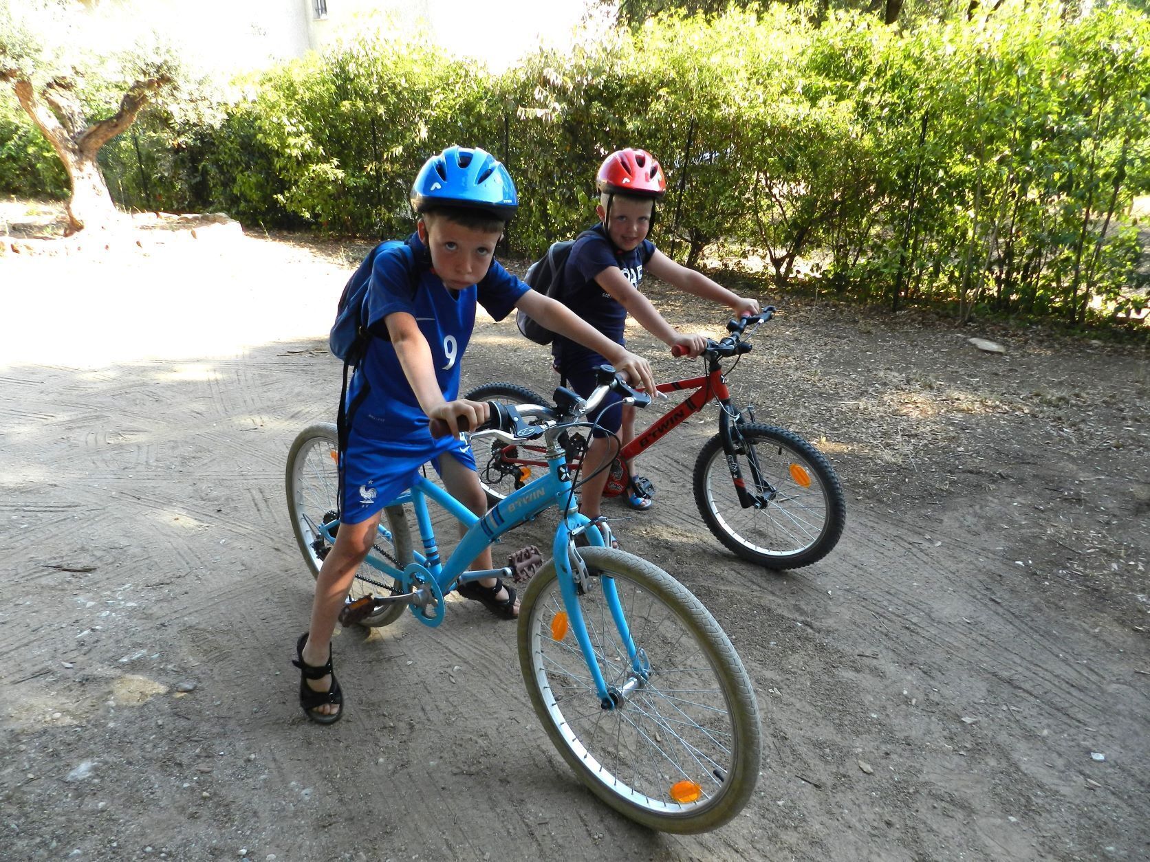 Two children riding bicycles on a dirt path with greenery in the background.
