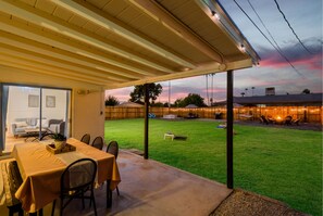 Un patio couvert avec une table et des chaises, une pelouse et un bâtiment dont on aperçoit des places assises à l’extérieur à l’arrière-plan.