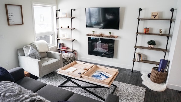 Uma sala de estar moderna com um sofá cinzento, uma mesa de centro, uma lareira e uma TV fixada na parede.