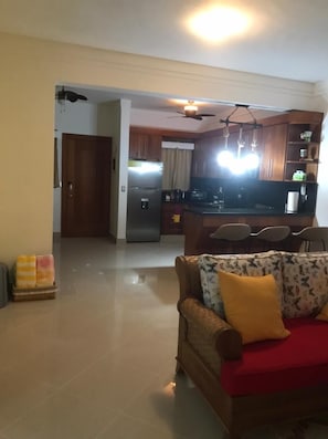 A living room with a sofa and a television. There is a kitchen in the background with wooden cabinets and a refrigerator.