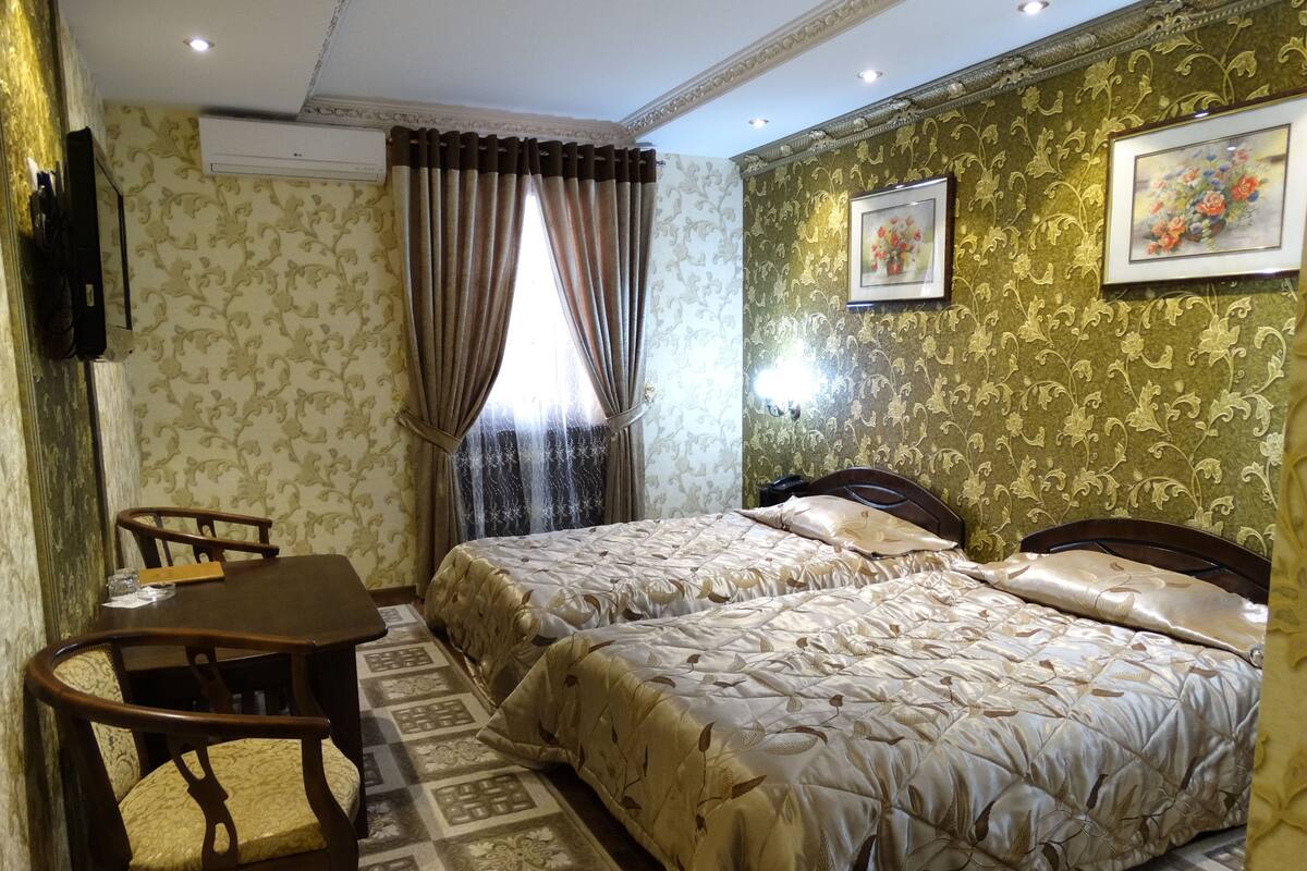 A hotel room with two beds, a wooden chair, a small table, and a window with curtains.