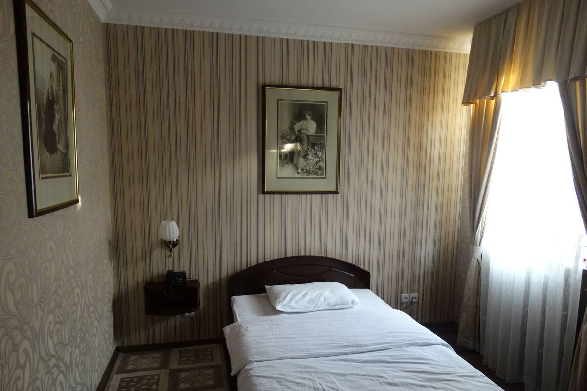 A hotel room with a bed, a nightstand with a lamp, a framed picture on the wall, and a window with curtains.
