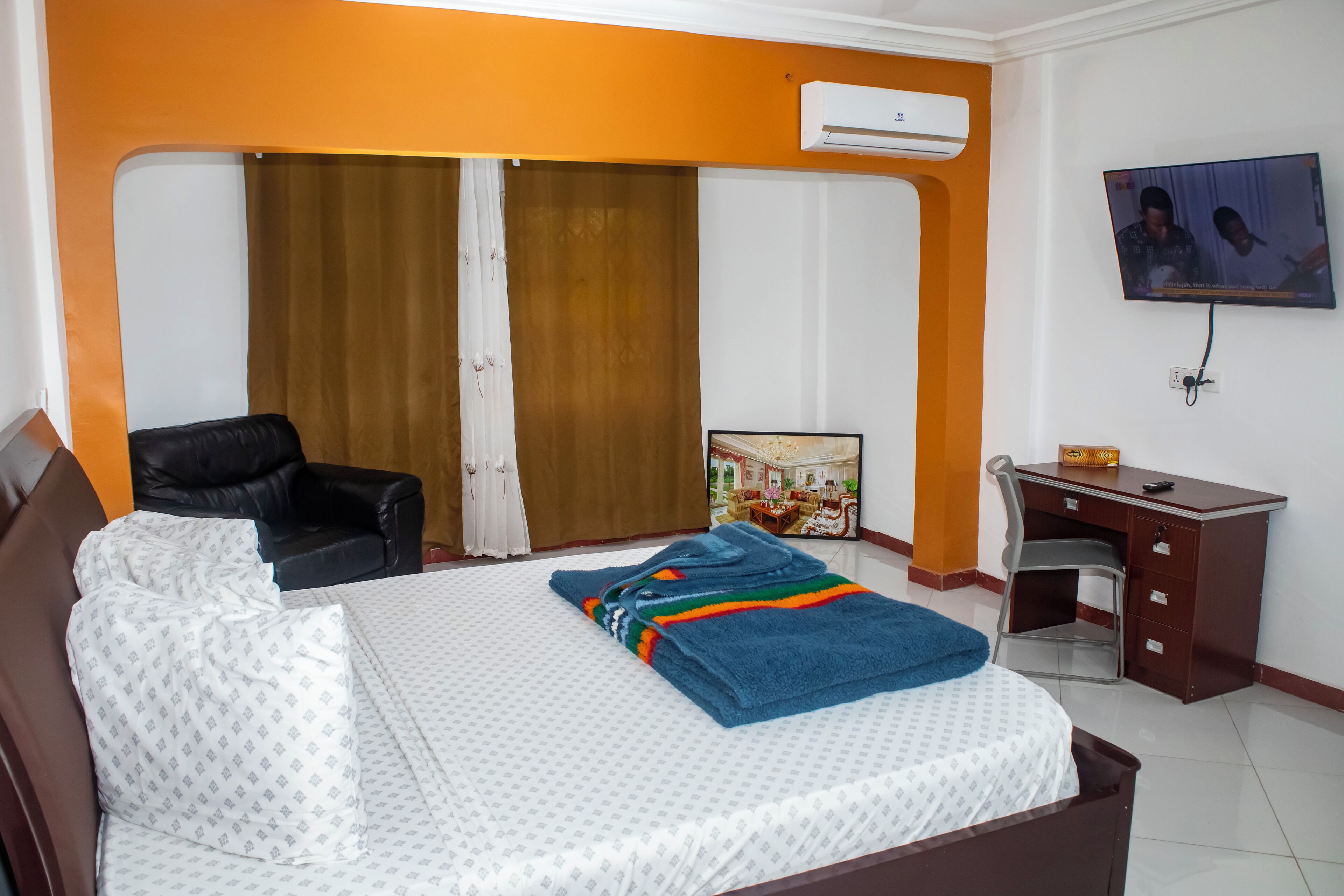 A hotel room with a bed, a desk, a chair, a TV, and an air conditioning unit.