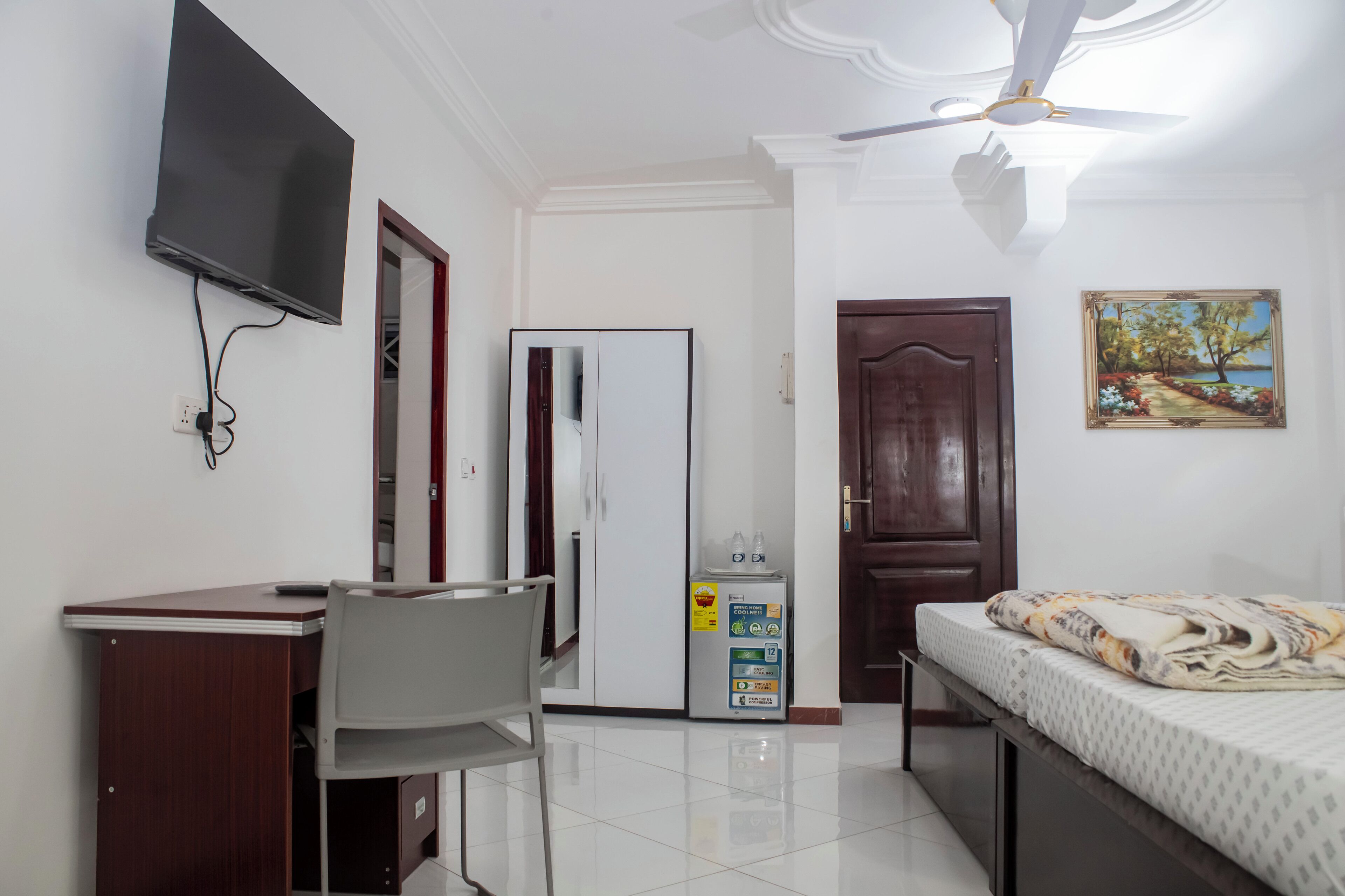 A bedroom with a bed, a desk with a chair, a wardrobe, a television mounted on the wall, a ceiling fan, a door, and a painting on the wall.
