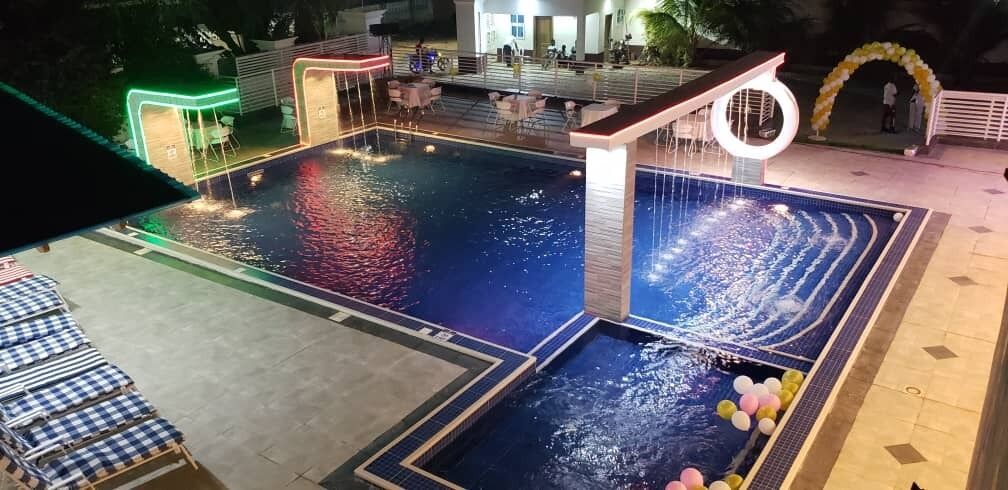 Una piscina con múltiples elementos acuáticos, incluyendo un tobogán y una fuente. Hay sillas de descanso y mesas alrededor de la zona de la piscina.
