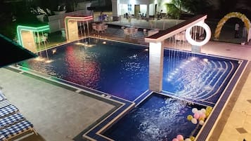 Una piscina con múltiples elementos acuáticos, incluyendo un tobogán y una fuente. Hay sillas de descanso y mesas alrededor de la zona de la piscina.