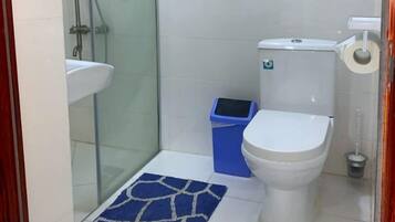 A small bathroom with a glass shower enclosure, a white toilet, a blue trash bin, and a white sink.