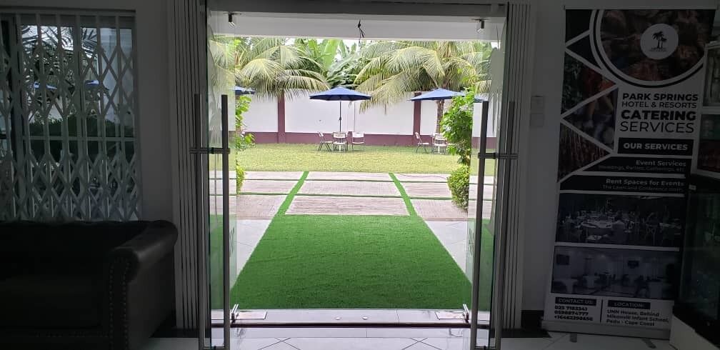 Entrada de un hotel con puerta de vidrio, vista a un patio con palmeras y césped verde, y un cartel publicitario de servicios de catering de Park Springs Hotel & Resorts.