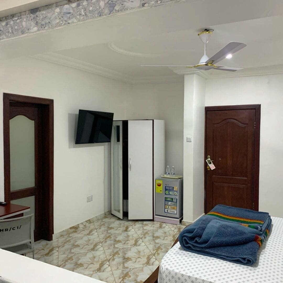 A bedroom with a bed, a wardrobe, a television, a ceiling fan, and a small refrigerator.