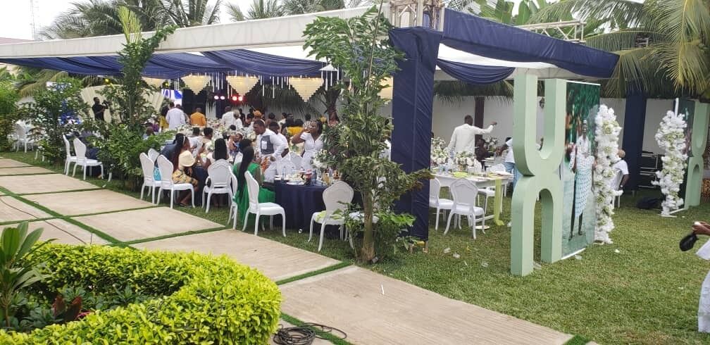 Un evento al aire libre con mesas y sillas redondas, rodeado de follaje y elementos decorativos.