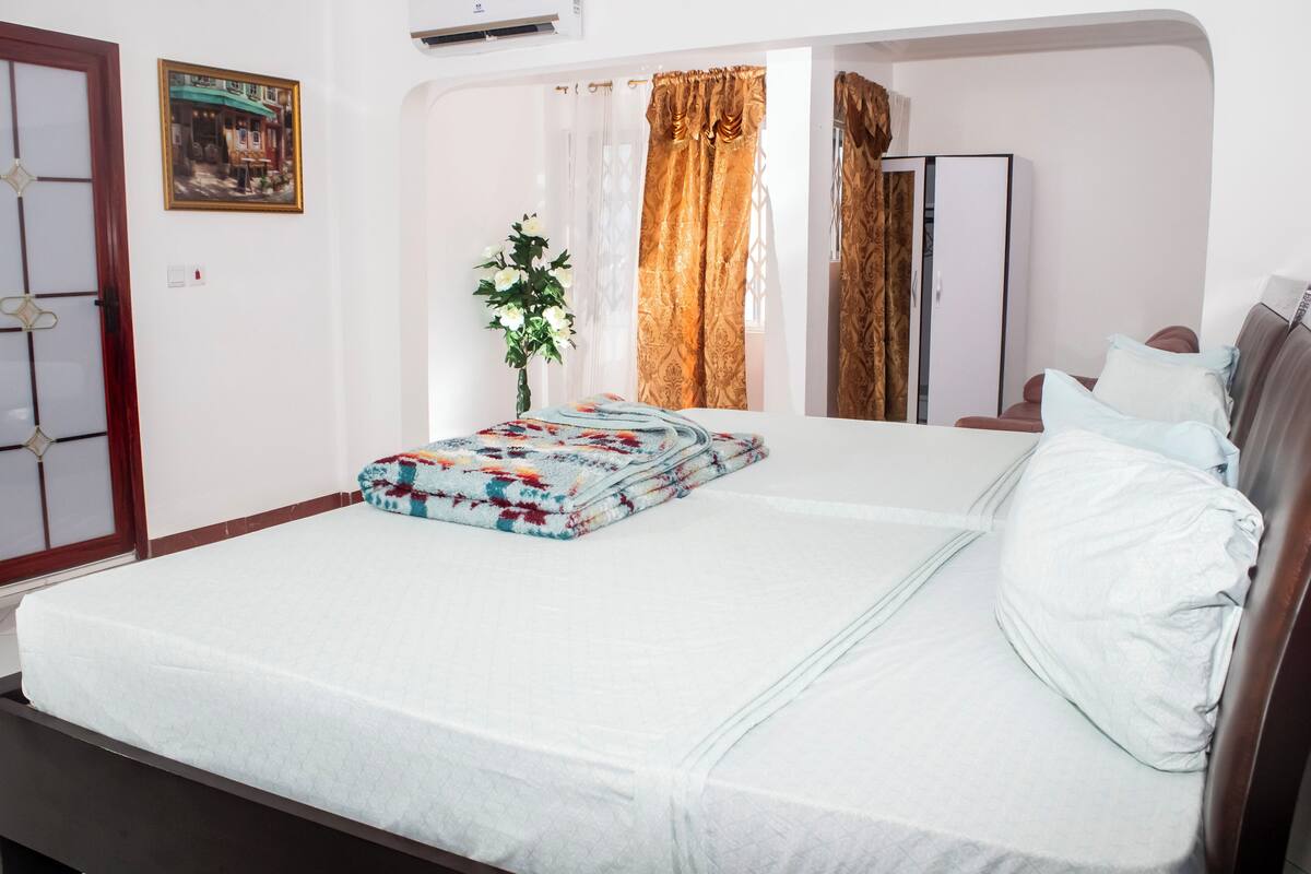 A neatly made bed with a floral-patterned blanket and pillows, a vase with flowers, and a painting on the wall.