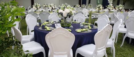 Un evento formal al aire libre con mesas redondas cubiertas con manteles azul oscuro, sillas blancas y centros de mesa florales.