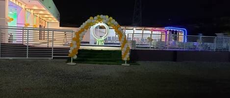 Un espacio para eventos al aire libre con escenario, decorado con un arco floral e iluminado por luces de escenario.