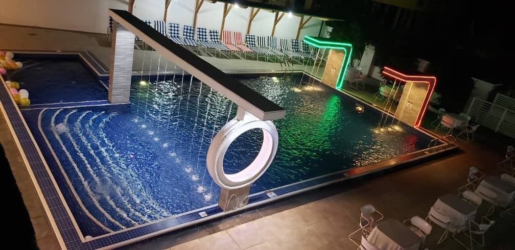 Una piscina interior con un elemento acuático circular, sillas de descanso y una zona junto a la piscina con una estructura iluminada con neón.