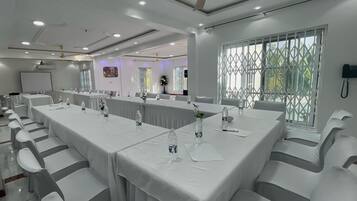 Una sala conferenze con lunghe tavolate, sedie bianche, ventilatori a soffitto e finestre con persiane.
