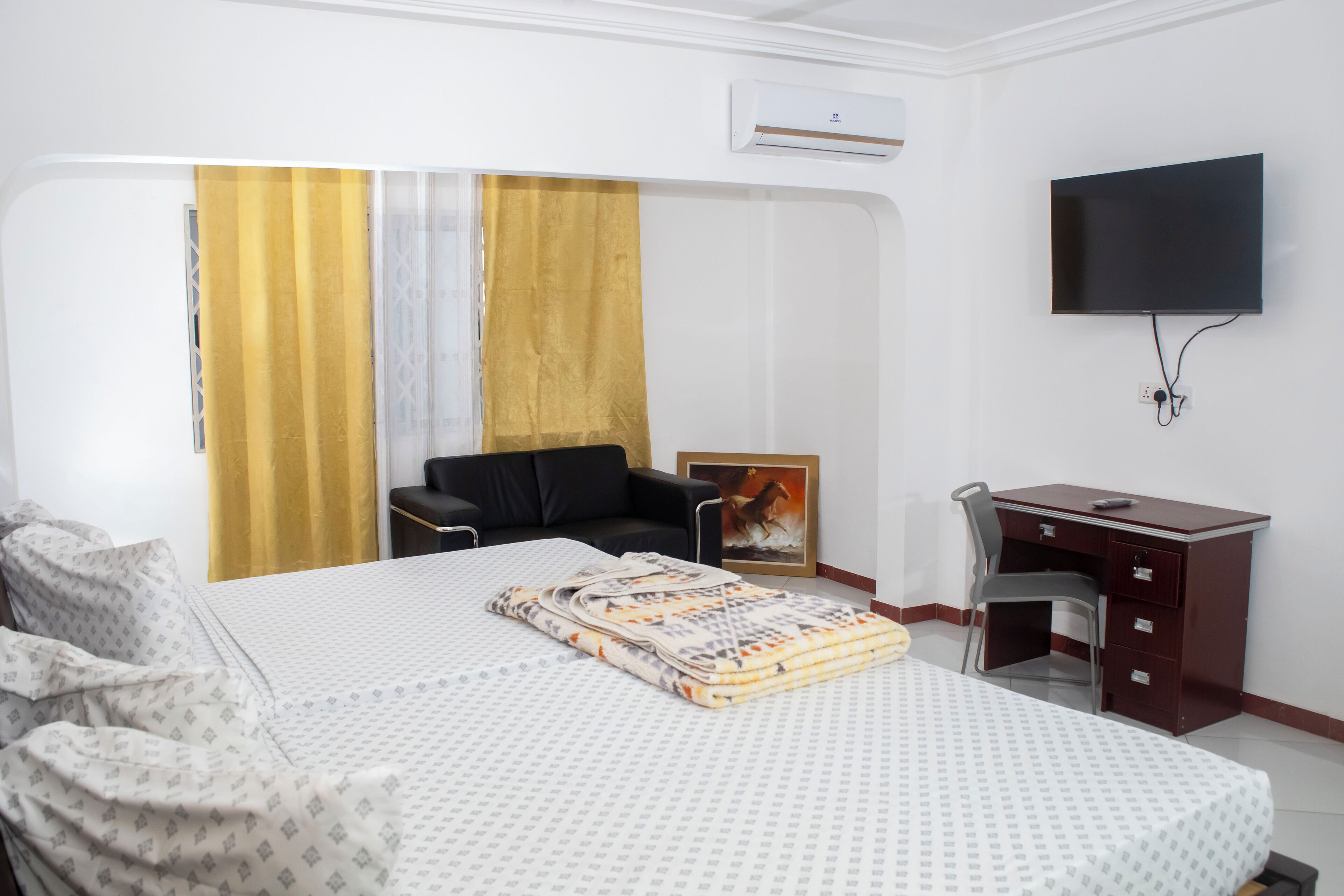 A hotel room with a bed, a desk, a chair, a TV, and an air conditioning unit.