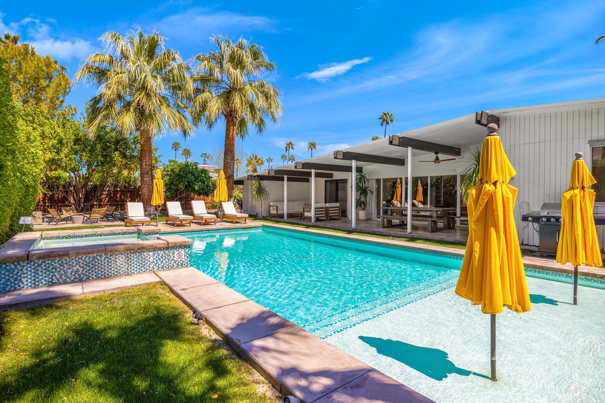 Une piscine entourée de palmiers, agrémentée de chaises longues et de parasols jaunes, et bordée d’une terrasse couverte.