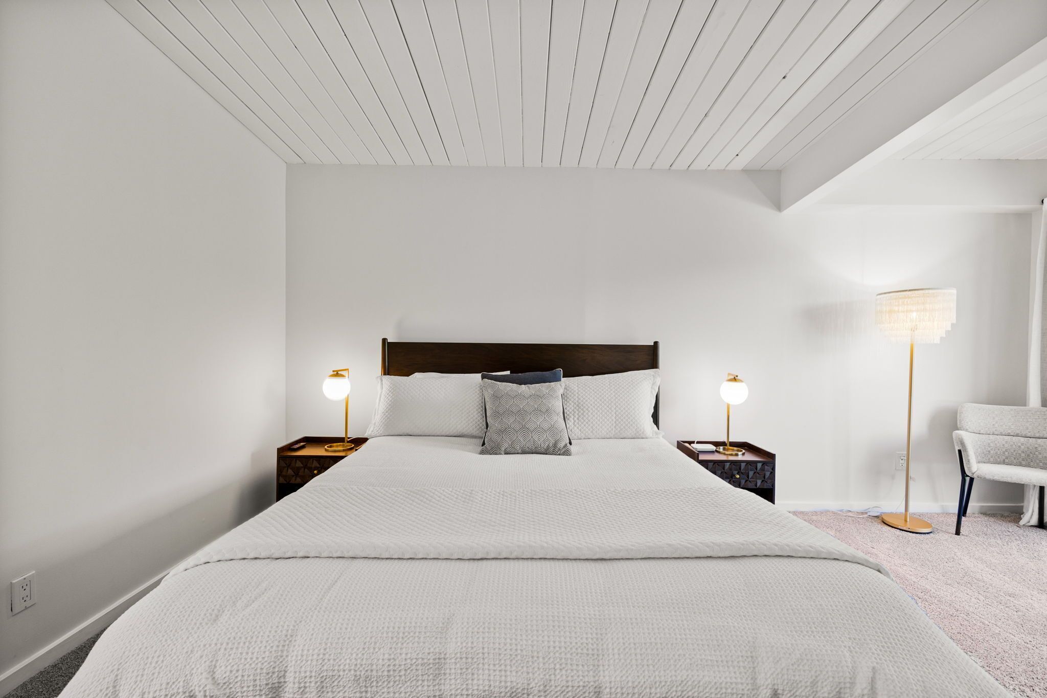 Un dormitorio ordenado con una cama grande, dos mesitas de noche con lámparas, una silla y una lámpara de pie.