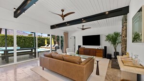 En moderne stue med en stor lædersofa i brunt, et glasbord og et fladskærms-tv. Der er en væg med detaljer i sten, og et stort vindue med udsigt over en pool.