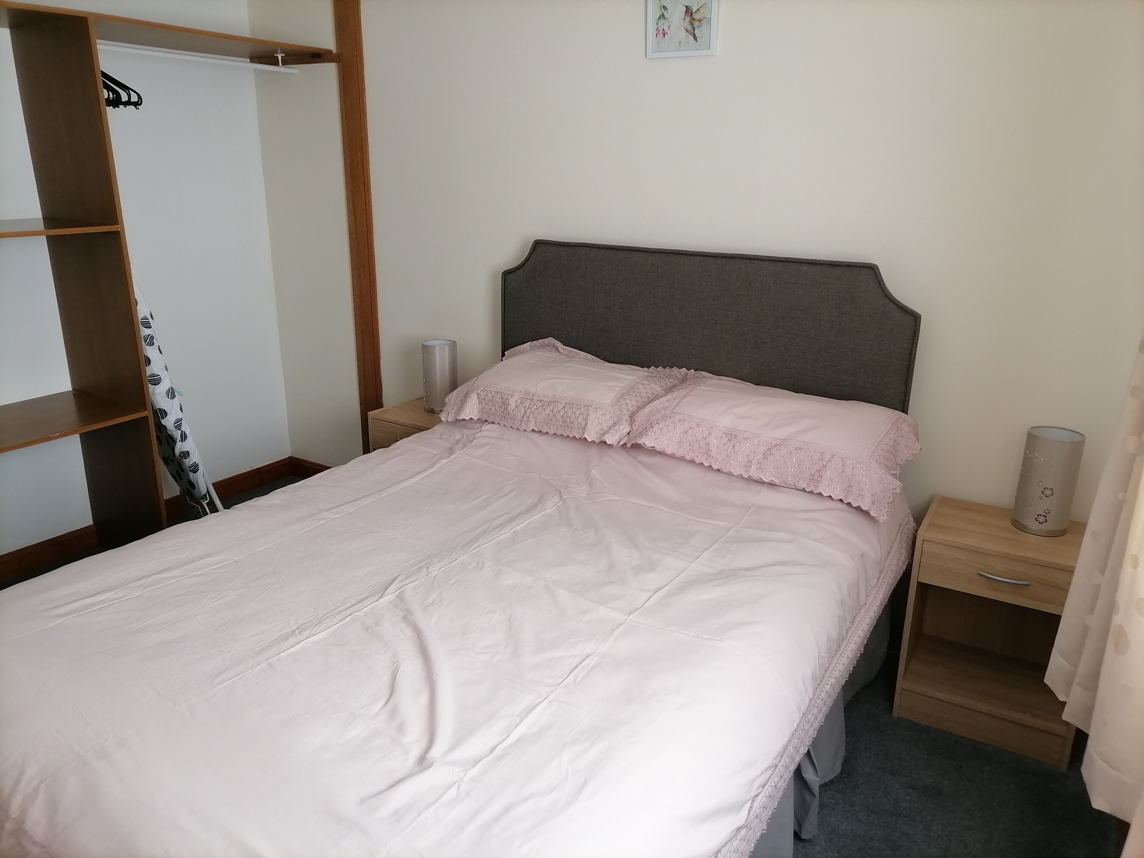 En snyggt bäddad säng med ett rosa överkast och kuddar, ett träbord vid sängen med en lampa och en garderob med en klädstång.