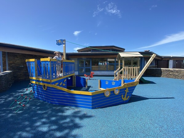 Un espace de jeux pour enfants avec une structure de jeu en forme de bateau bleu et un bâtiment en arrière-plan.