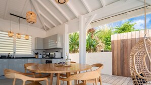 A modern kitchen with white cabinets, a dining area with wooden chairs and a round table, and a view of a wooden fence and greenery outside. - Villa Rackam, balcony on the Baie des Saintes with pool and 300m from beaches (Terre-de-Haut)