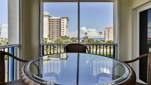 A balcony with a glass table and wicker chairs, overlooking a coastal view with buildings and palm trees.