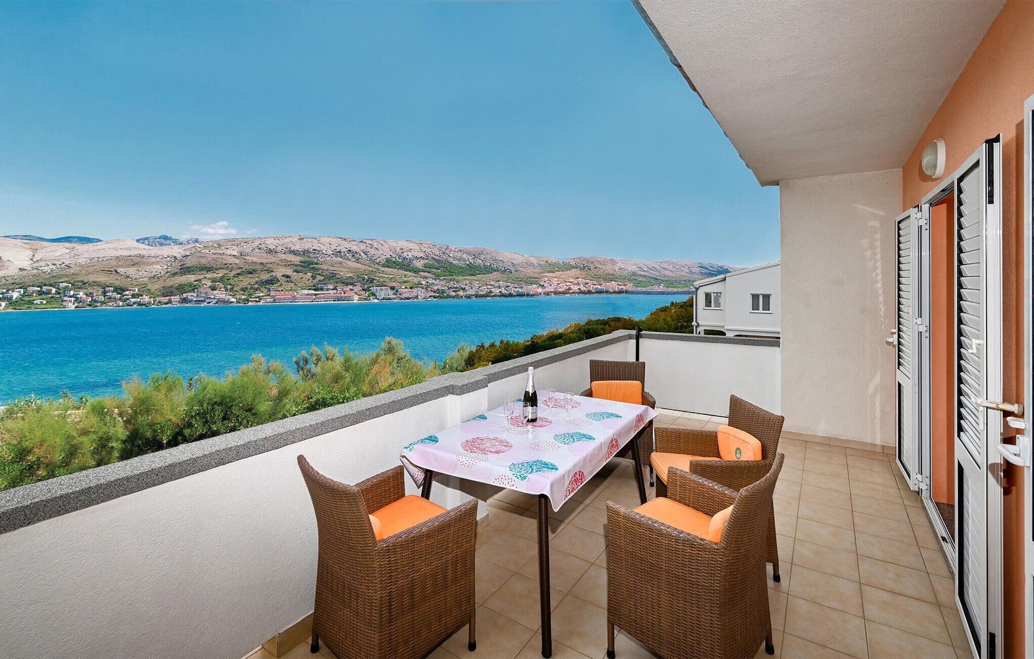 A balcony with a table and chairs, overlooking a coastal view with a clear sky.