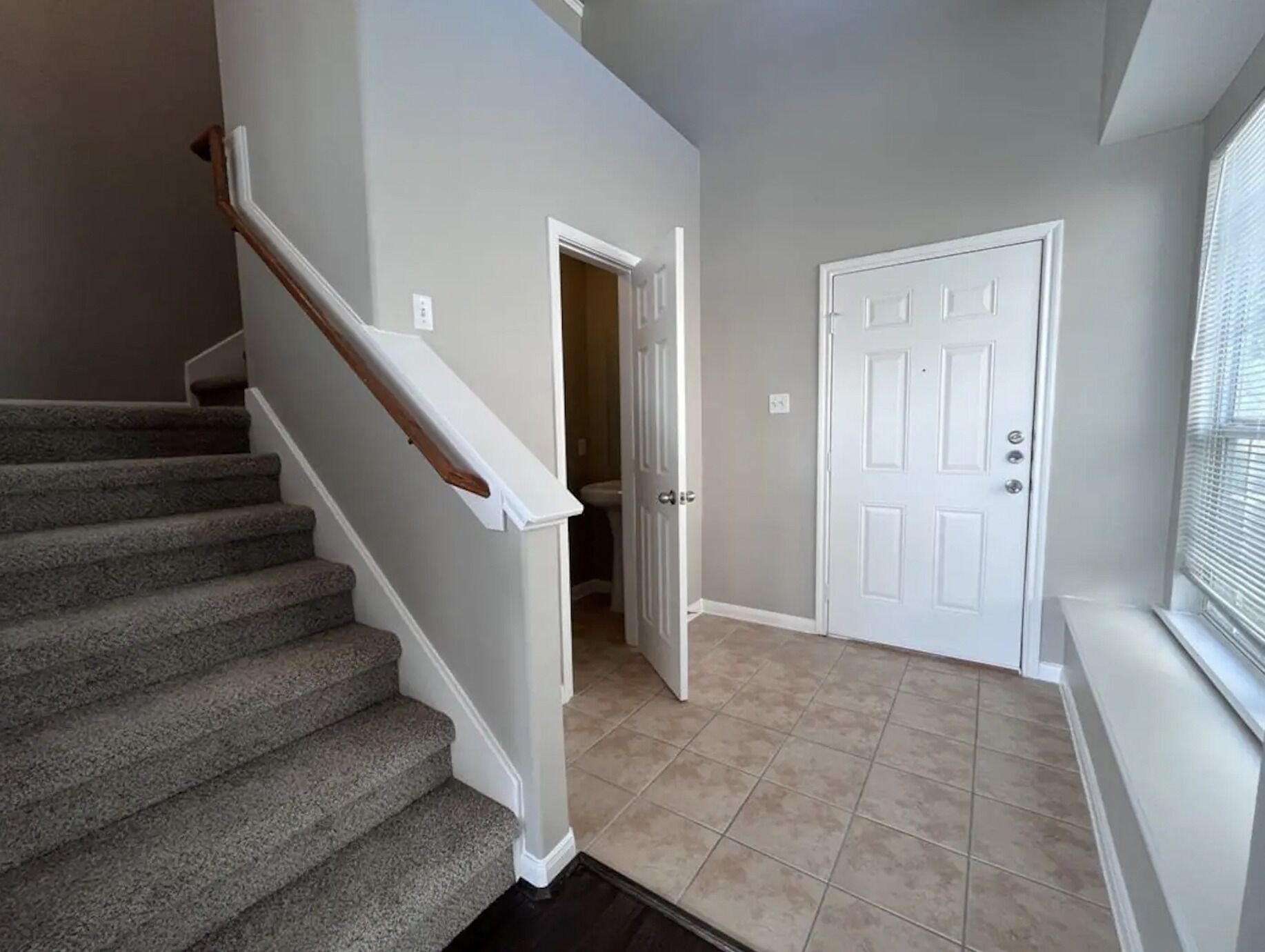 Un escalier avec des marches recouvertes de moquette et une rampe en bois, menant à une pièce dotée d’une porte blanche et d’un sol carrelé de couleur beige.