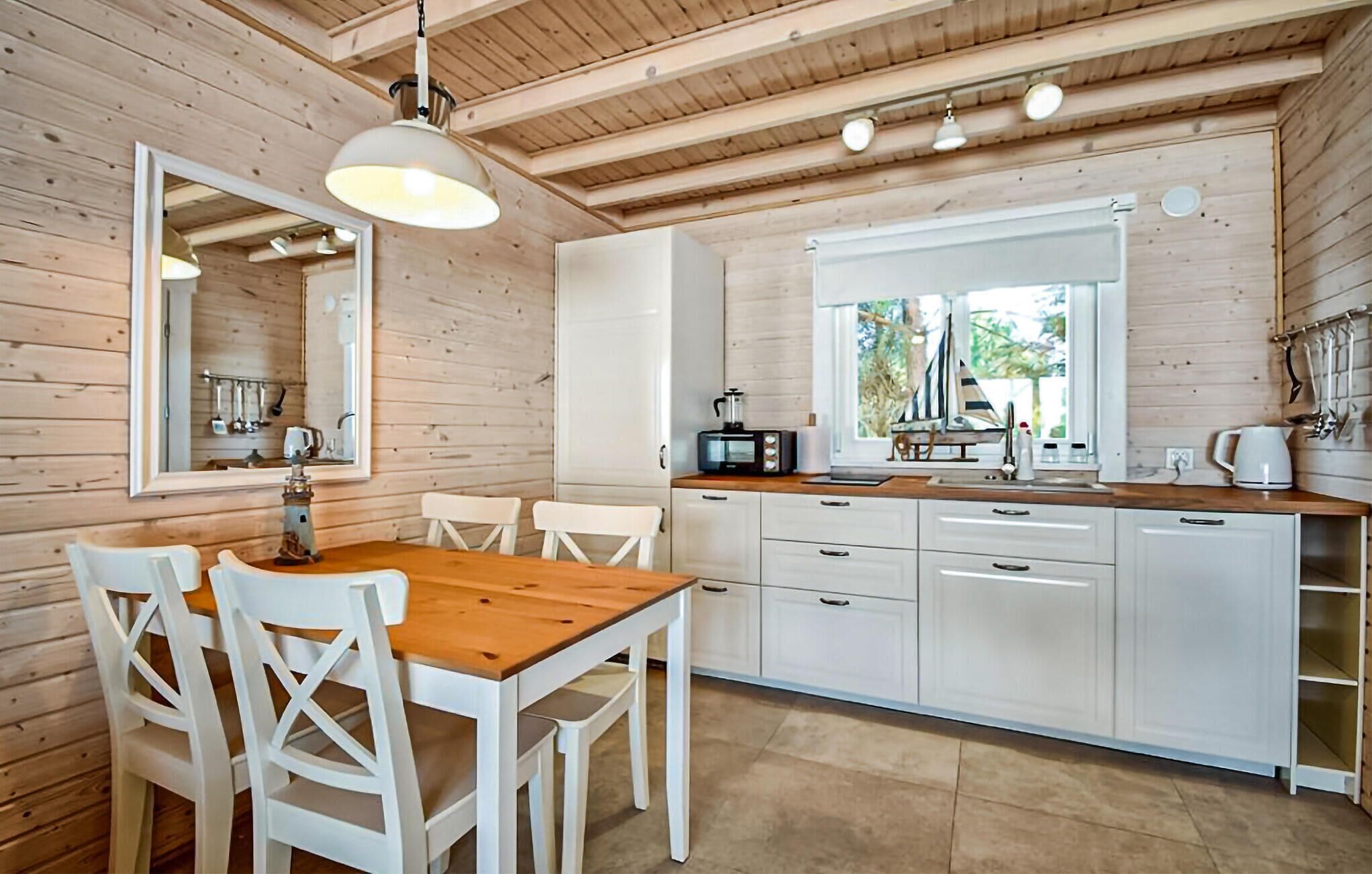 Interior de uma cabana de madeira com uma mesa de jantar, cadeiras brancas, cozinha com armários brancos, micro-ondas e janela com vista para o verde.