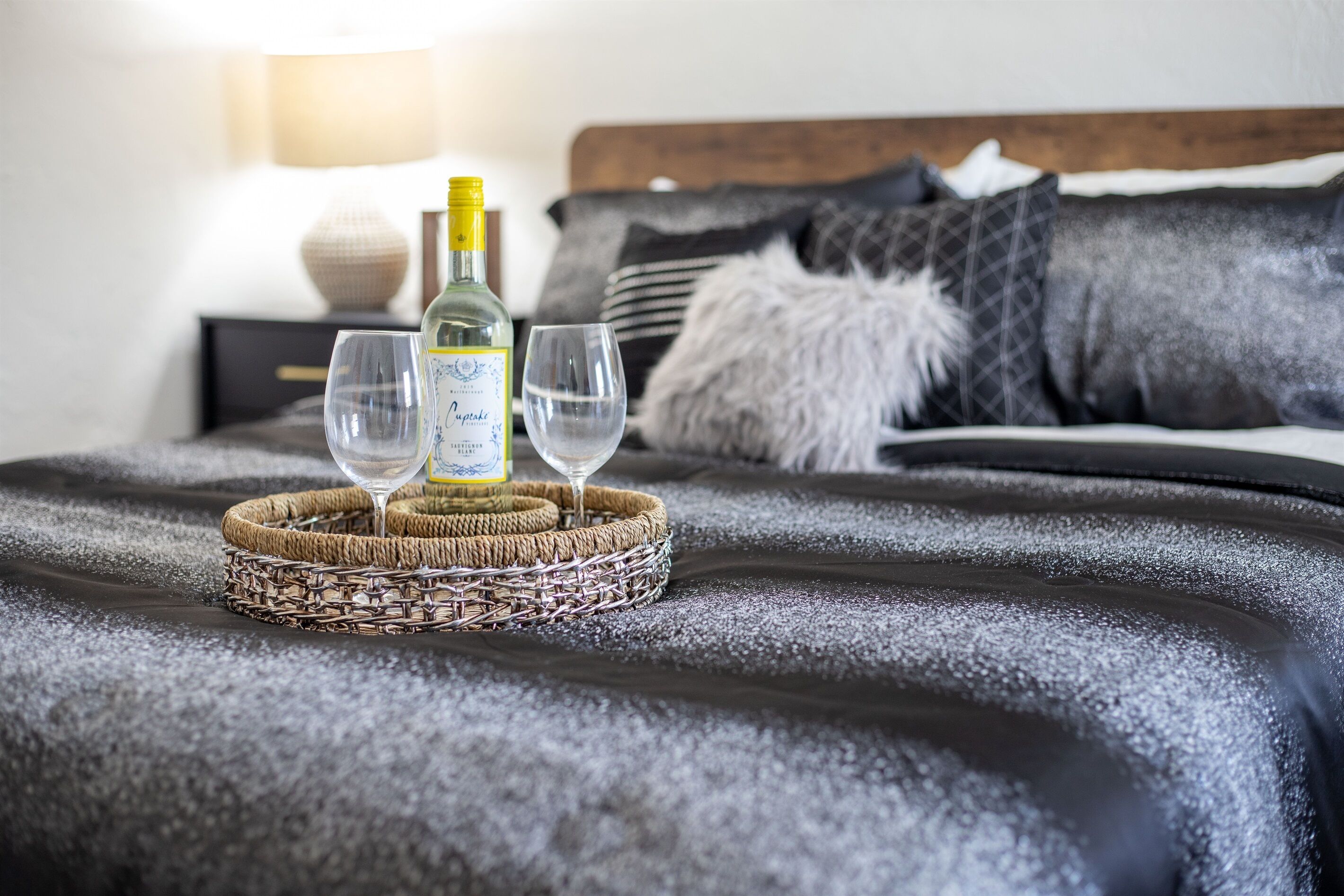 Un letto con una coperta scura e materassata, una bottiglia di vino e due bicchieri su un vassoio di vimini.