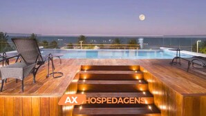 Un área de piscina en la azotea con terraza de madera, sillones de descanso y vista al océano al atardecer.