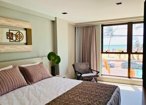 Quarto de hotel com uma cama grande, uma cadeira, uma janela com vista para a praia e uma obra de arte na parede.