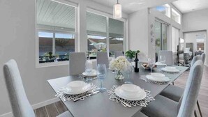 A modern dining area with a large table set for six, large windows, and a view of the outdoors. - Fun gameroom,big yard,pool+spa,fire pit (Desert Hot Springs)