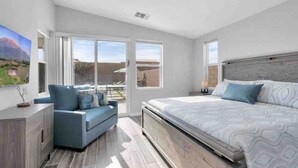 A modern bedroom with a large bed, a blue sofa, and a view of the outdoors through large windows. - Fun gameroom,big yard,pool+spa,fire pit (Desert Hot Springs)
