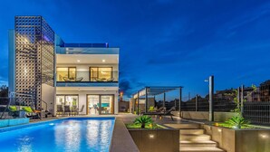 Et moderne hus med to etager, swimmingpool, udendørs møbler og en oplyst balkon.