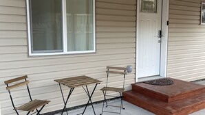 A covered porch with a table and two chairs, a white door, and a small set of steps leading to a fire pit.
