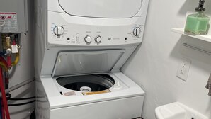 A compact washer and dryer unit with control knobs, a safety warning label, and a nearby sink with a towel rack.