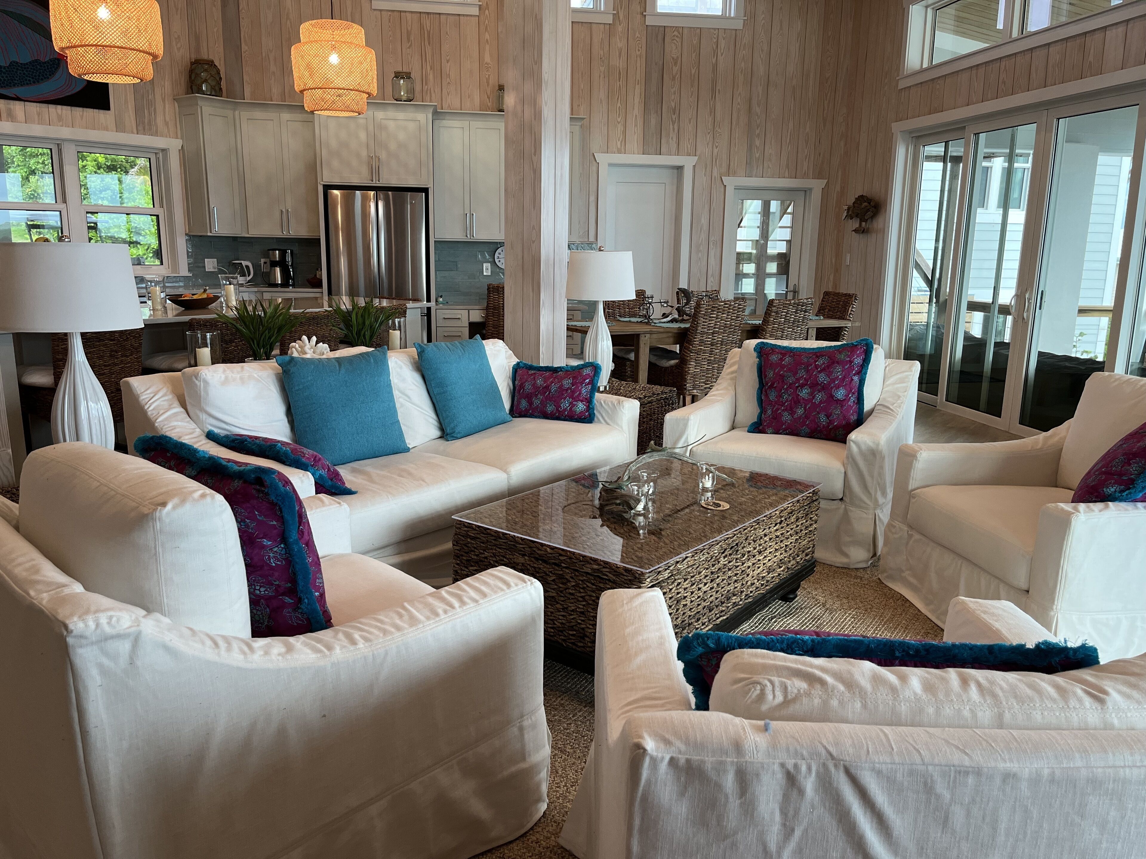 A spacious living room with white sofas and armchairs, a glass coffee table, and a kitchen in the background.