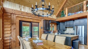 A log cabin interior with a wooden dining table, wicker chairs, a chandelier, a kitchen with dark cabinets and stainless steel appliances, and a balcony with a railing. - Chalet Belle Cordélia with Spa and AC (Nominingue)