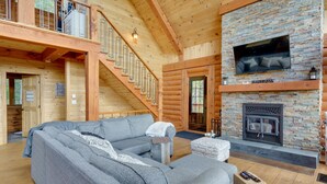 A log cabin interior with a gray sectional sofa, a stone fireplace, and a wooden staircase. - Chalet Belle Cordélia with Spa and AC (Nominingue)