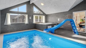 Une piscine intérieure avec un toboggan bleu, de grandes fenêtres et une vue sur une maison à l’extérieur.