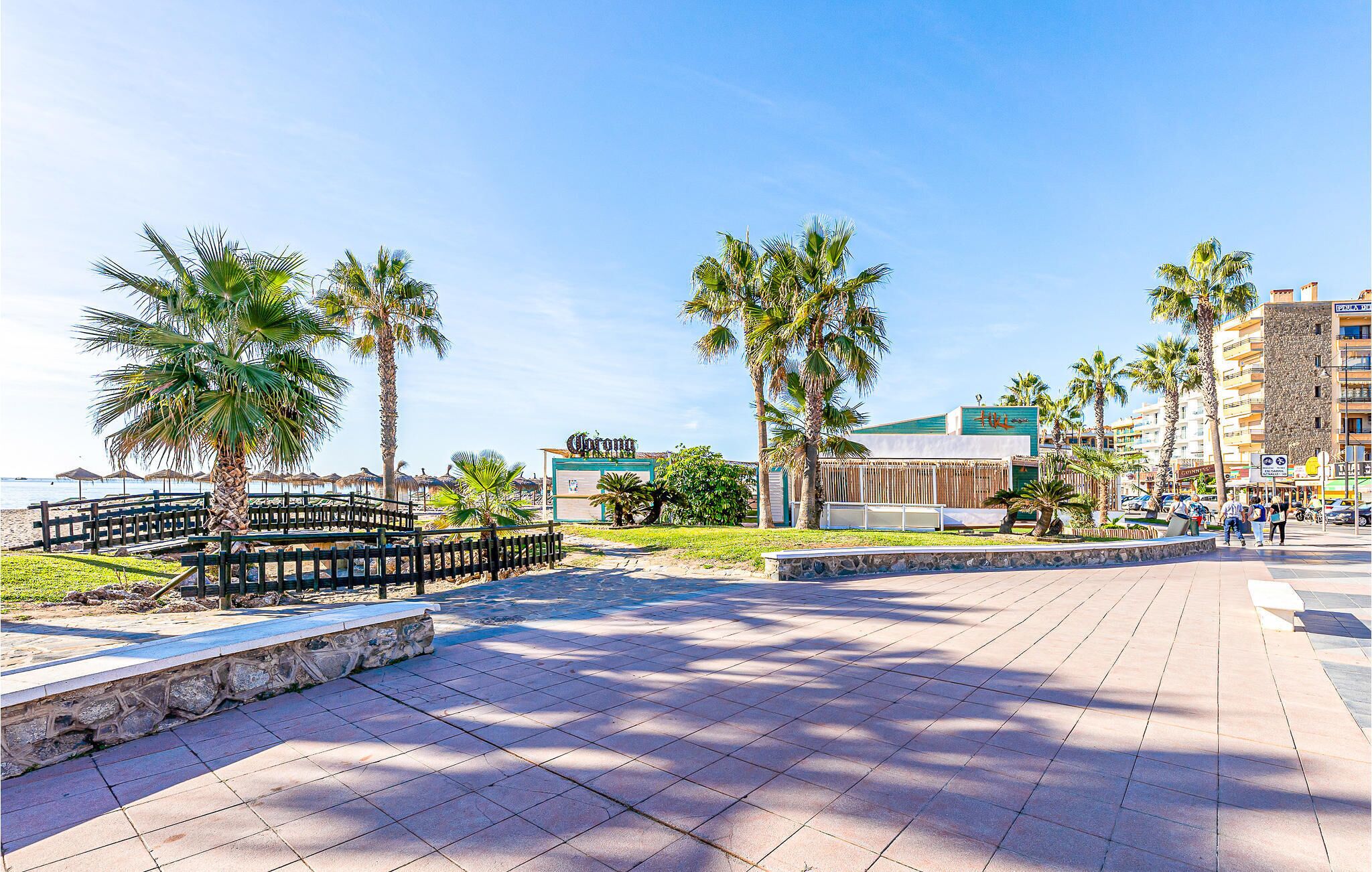 A paved promenade with palm trees, a beach in the background, and a building with a sign.