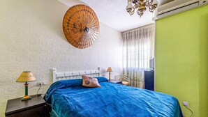 A bedroom with a bed, two bedside tables, a lamp, a ceiling fan, and a large wall hanging.