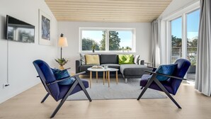Ein modernes Wohnzimmer mit einer Couch, zwei Sesseln, einem Couchtisch und einer Stehlampe. Ein großes Fenster bietet Blick auf Bäume und einen Balkon.