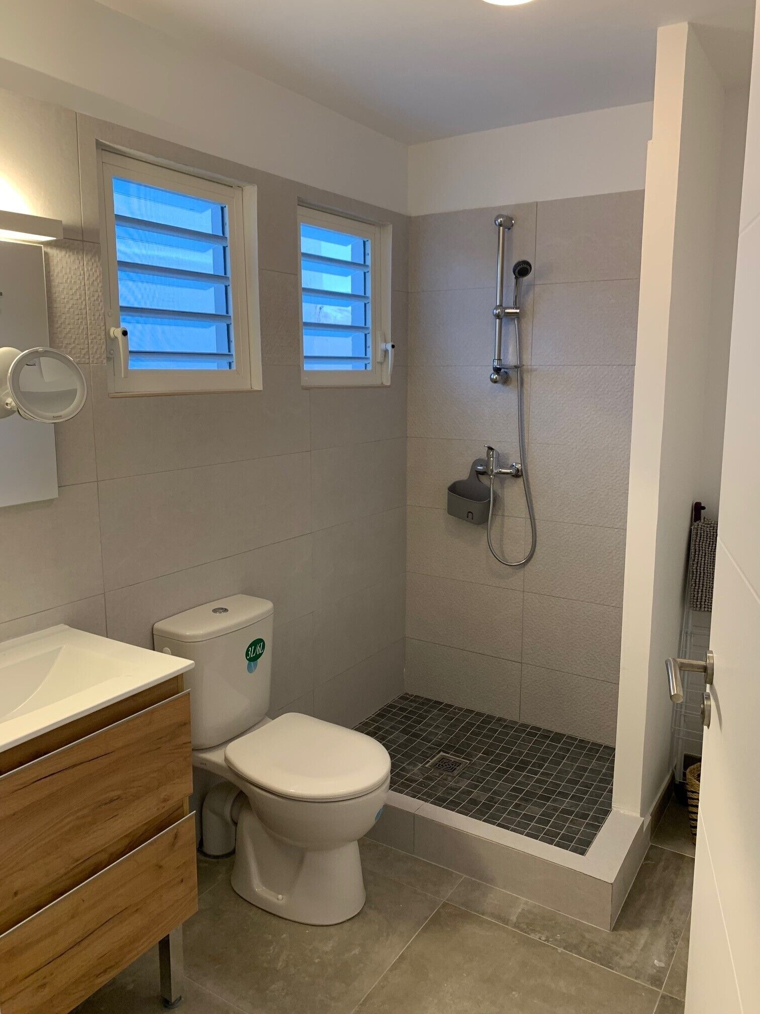 A modern bathroom with a toilet, bidet, and shower. There are two windows with blinds, a mirror, and a sink with a wooden vanity.
