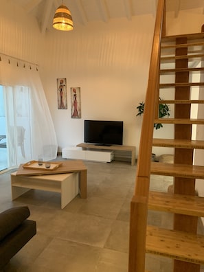 Ein modernes Wohnzimmer mit einer Holztreppe, einem Flachbildfernseher, einem Glastisch und einem großen Fenster mit durchsichtigen Vorhängen.