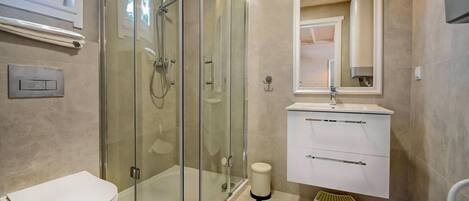 A modern bathroom with a glass-enclosed shower, a white toilet, a vanity with a mirror, and a small trash bin.