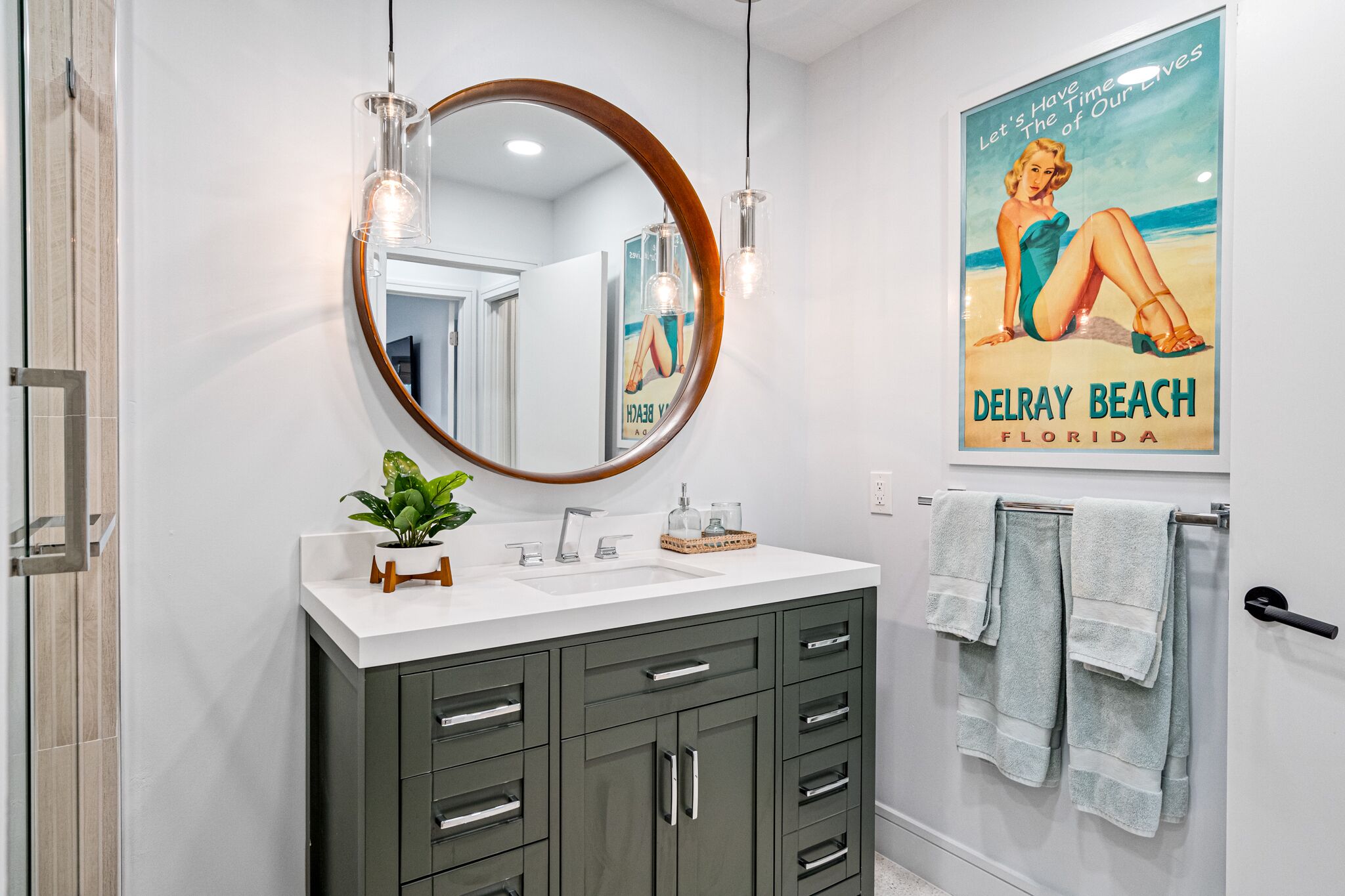 Een badkamer met een ronde spiegel, een witte wastafel, donkere kastjes, een potplant en een poster van Delray Beach, Florida.